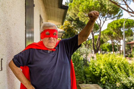 Funny and happy senior man posing as superhero with red cape and mask is raising arm preparing to fly in a quiet residential neighborhoodの写真素材
