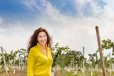 Yellow dressed menopausal woman with wrinkles and eye bags enjoying nature of a countryside while walking in the kiwi orchard in Autumn seasonの写真素材