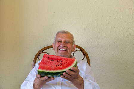 Happy old man holding slice of watermelon over yellow wall backgroundの写真素材