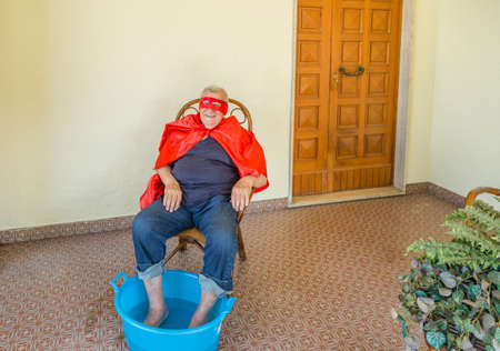 elderly man wearing a superhero costume and sitting on wicker chair on yellow background while having footbath in water basin in home patioの写真素材