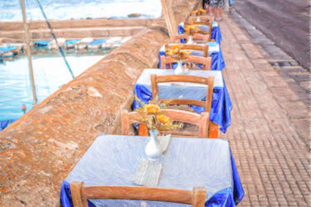 Dinner tables in Italian restaurant on brown stone parapet on the sea bay of Gallipoli (Le) in Italy in a summer sunny day: plastic fake yellow flowers with large petals and green leaves in white vase, White and blue gingham napkins and tablecloth  on eleの写真素材