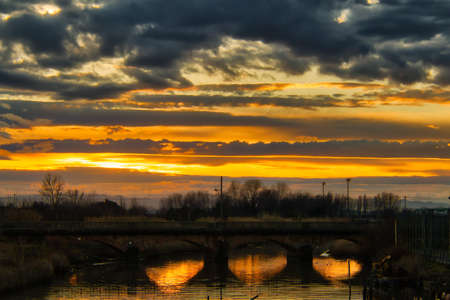 Rays of sun through clouds at sunset reflected on the waters of a river passing under the arches of...