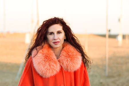 Menopausal woman with wrinkles dressed with orange fur collar and coat on winter beachの写真素材