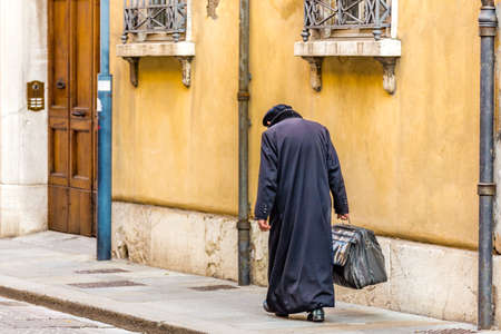 elderly man with black dress and hunched back walking in a town street with a heavy duffel bagの写真素材