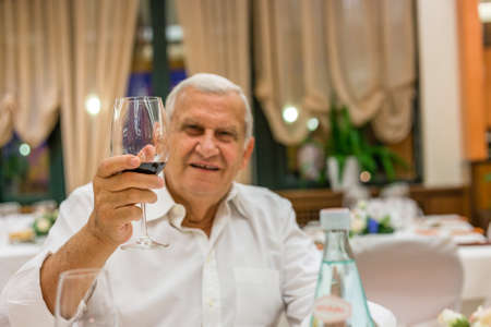 Old man is holding a glass of red wine while having dinner at Italian restaurantのeditorial素材