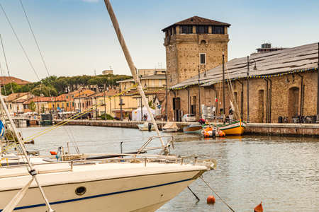 CERVIA (RA), ITALY - MAY 17, 2019: boats mooring in port channel of Cervia with medieval tower in...