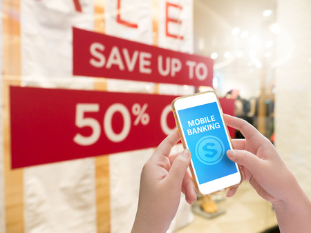 Hand holding mobile phone with Mobile banking word with blur save up to 50% shop window displayo...