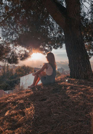 Blonde-haired woman sitting in a field in the shade of a tree looking for relaxation at sunset at the top of the mountain.の写真素材