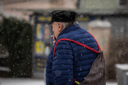 Prague, Czech Republic. 01-12-2021. Old man smoking while walking on a snowing day with mask during covid-19 from hradcanska tram station at Prague 6のeditorial素材