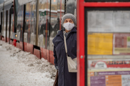Prague, Czech Republic. 02-08-2021. Old woman with mask and walking stick is looking at the tram schedule in the city center of Prague on a cold winter snowy day.のeditorial素材