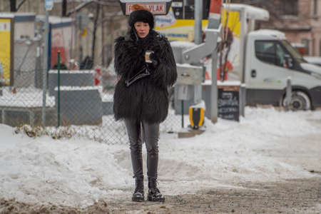 Prague, Czech Republic. 02-08-2021. Elegant woman dress in black holding a coffee cup waiting green light to cross the street in the city center of Prague on a cold winter snowy day.のeditorial素材