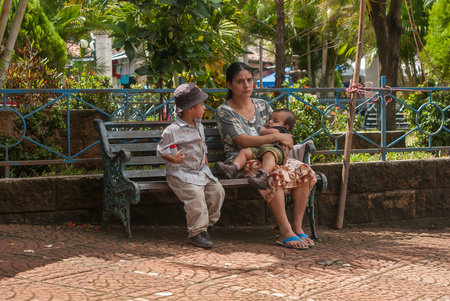 Suchitoto, El Salvador. 03-21-2019. Sad mother with two children sitting on a bench at the sunday market in the old city of Suchitoto.のeditorial素材