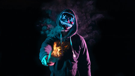 Man in a hood with a skull on his face holding a burning fire in his handsの写真素材