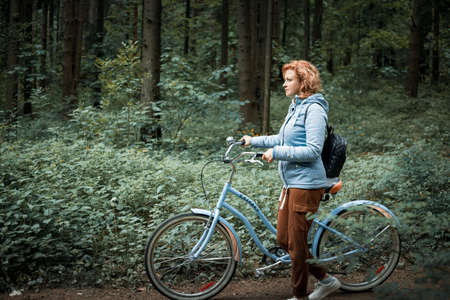 A blonde woman in a blue jacket walks through the forest with a blue bicycleの写真素材
