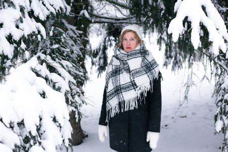 A blonde woman in winter stands under a fir-tree in the snow.