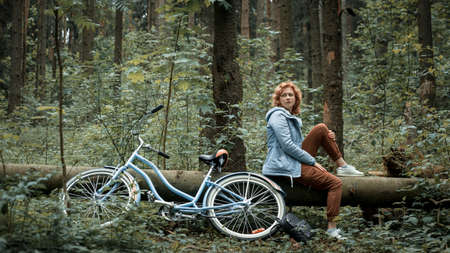Woman with blue bike sitting on a fallen tree in the forestの写真素材