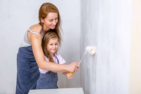 Repair in the apartment. Happy family mother and daughter in aprons paint the wall with white paint. Mother helps her daughter to paint the wall with a rollerの写真素材