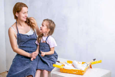 Repair in the apartment. Happy family mother and daughter in aprons paint the wall with white paint. Mother and daughter sit side by side and smear each other with paint and laughの写真素材