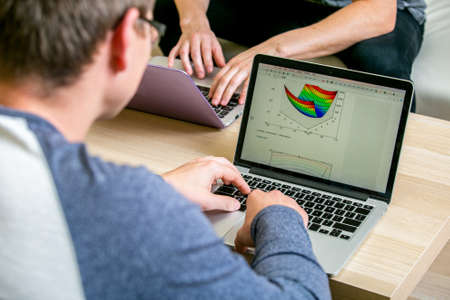 Two young people working on laptops in the office, writing a program, correct the text. Sit at the table opposite each other. A man is viewing a chart on the display.の写真素材