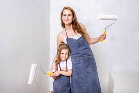 Repair in the apartment. Happy family mother and little daughter in blue aprons paints the wall with white paint. Mom and daughter are holding paint rollers and smiling. Horizontal portraitの写真素材