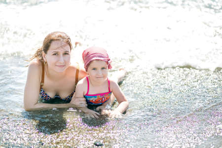 Beautiful young mother and daughter having fun resting on the sea. They lie on the sand in the water and looking at the camera. Horizontal photoの写真素材