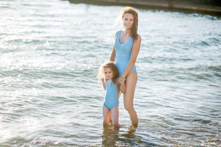 Beautiful young mother and daughter having fun resting on the sea. They stand in the water in the same swimsuit and smile. Horizontal photoの写真素材