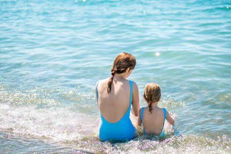 Beautiful young mother and daughter having fun resting on the sea. They sit in the water in the same swimsuit, their backs in the frame. Horizontal photoの写真素材
