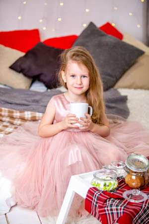 Pretty little girl 4 years old in a pink dress. Child in the Christmas room with a bed, eating candy, chocolate, cookies and drinking tea, feeding teddy bears and laughing, good mood.の写真素材