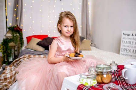 Pretty little girl 4 years old in a pink dress. Child in the Christmas room with a bed, eating candy, chocolate, cookies and drinking tea, feeding teddy bears and laughing, good mood.の写真素材