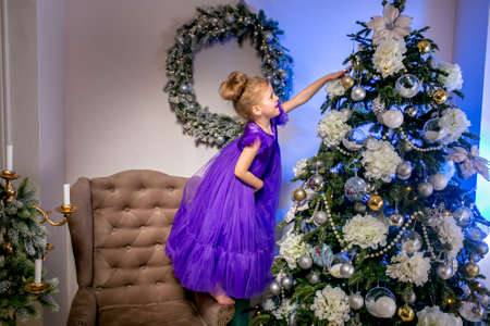Pretty little girl 4 years old in a blue dress. Baby in Christmas room with teddybear, big clock, christmas tree, brown armchair, candles and gifts.の写真素材