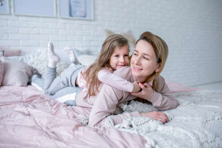 Beautiful young mother and her little daughter are lying together on the bed in the bedroom,...