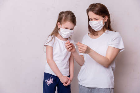 The concept of coronavirus quarantine. Young woman and her daughter in medical masks in isolation at home. Mom measures the temperature of her daughter. Theme of health and medicine.の写真素材