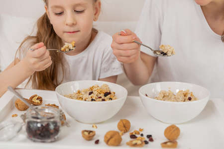 Happy mom and daughter have healthy breakfast on bed in a light bedroom on a sunny morning. Oatmeal porridge with nuts and raisins. Healthy food and family relationships concept. Good mood. Close-upの写真素材