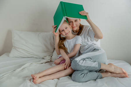 Happy young mother with daughter are reading a book in the light bedroom. They hold the book over their heads like a roof. Family relationships and home leisure. Good mood. Free spaceの写真素材