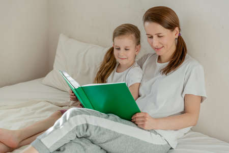 Happy young mother with daughter are reading a book in the light bedroom on a sunny morning. They look at the book together. Family relationships and home leisure. Good mood. Free space.の写真素材