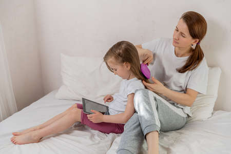 Happy young mother with daughter in the light bedroom. The daughter is engaged on the tablet while mom is combing her hair. Family relationships and home leisure. Good mood.の写真素材