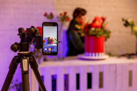 Florist workplace on the background of a white brick wall. A florist creates a flower arrangement in a red box and records an educational video on a smartphone. Online lesson, video conference.の写真素材