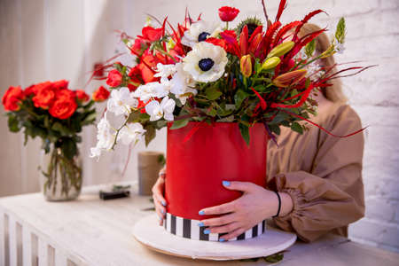 A flower arrangement in red shades of orchids, roses, sunflowers, lilies, alstroemerias and ornithogalums in a red box in the hands of a young florist girl. Professional florist work.の写真素材