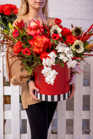 A flower arrangement in red shades of orchids, roses, sunflowers, lilies, alstroemerias and ornithogalums in a red box in the hands of a young florist girl. Professional florist work.の写真素材
