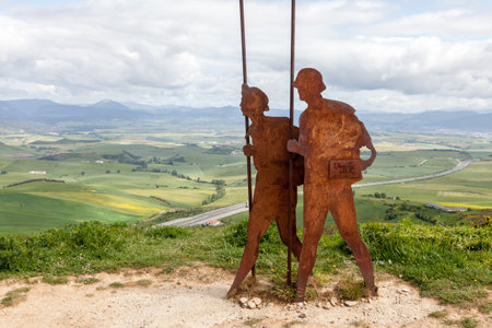 On the Camino de Santiago, metal sculptures of pilgrims walk toward stunning hills under a cloudy sky.の写真素材