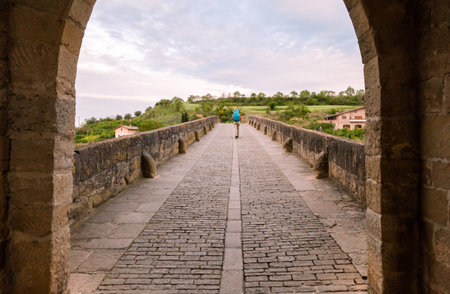 A person strolls on an ancient stone bridge in Obanos, Spain, enjoying the lush greenery and serenity of the landscape.の写真素材