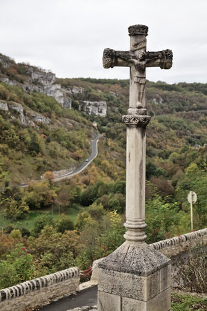 A stone cross stands at the edge of a cliff, overlooking the lush, colorful valley and winding road below in Rocamadour.の写真素材
