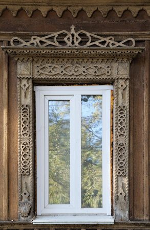 A white window is framed by elaborate carvings on a rustic building with greenery reflected.の写真素材