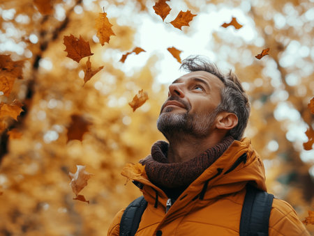 A man stands in a forest, gazing upward as orange leaves fall around him during the golden hour, enveloped in the season's beautyの素材