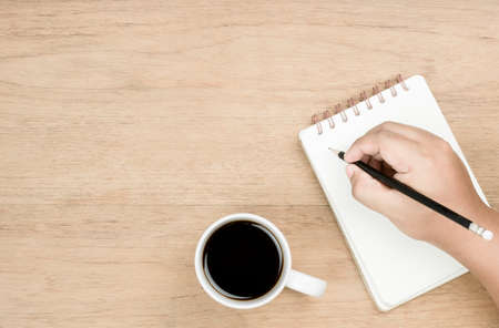 brown skin hand use black pencil writing on white page of small notebook and white coffee cup place...