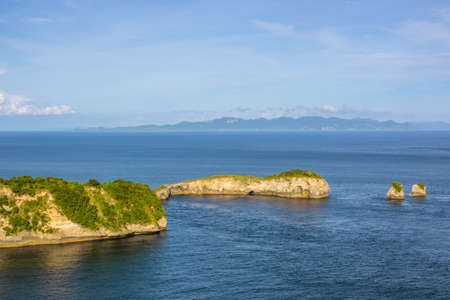 Indonesia. Strait between the islands. Day. Rocky coast with jungle and two small islets. Mountains and clouds on the horizonの写真素材