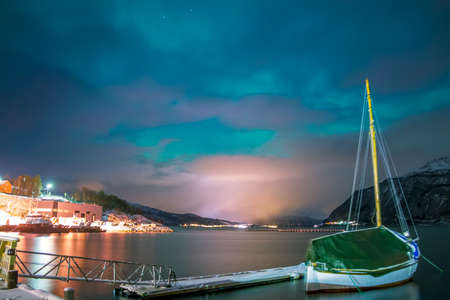 Night fjord in winter Norway. The boat is at the pier. The lights of the village on the horizon, surrounded by snow-capped mountains. Small northern lights in the skyの写真素材