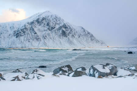 Winter Norway. Bay surrounded by snow-capped mountains. A ray of sunshine illuminates a lonely hut and part of the beachの写真素材