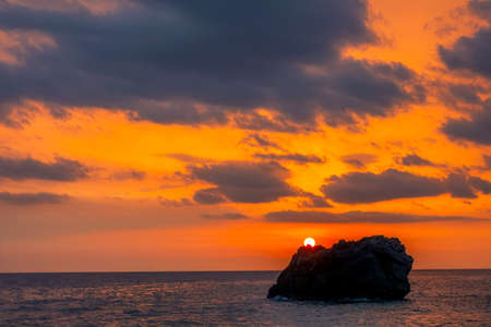 Sunset over a lonely rock in a calm sea. Amazing colors in the cloudy skyの写真素材