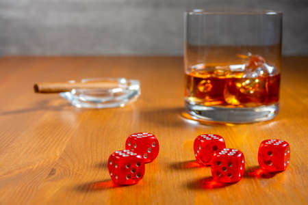 Red dice on a wooden table. Glass of whiskey with ice cubes and an ashtray with a cigar in the...